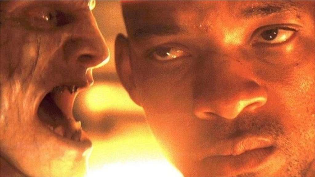 A close-up of two faces: on the left, a pale, screaming creature with sharp teeth; on the right, a man with a serious expression, illuminated by orange light.