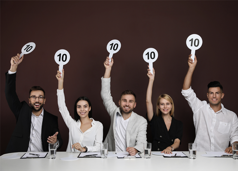 Five smiling judges sitting at a table hold up signs displaying the number 10. They appear pleased, and the background is a solid dark color. Each judge has a glass of water and papers in front of them.