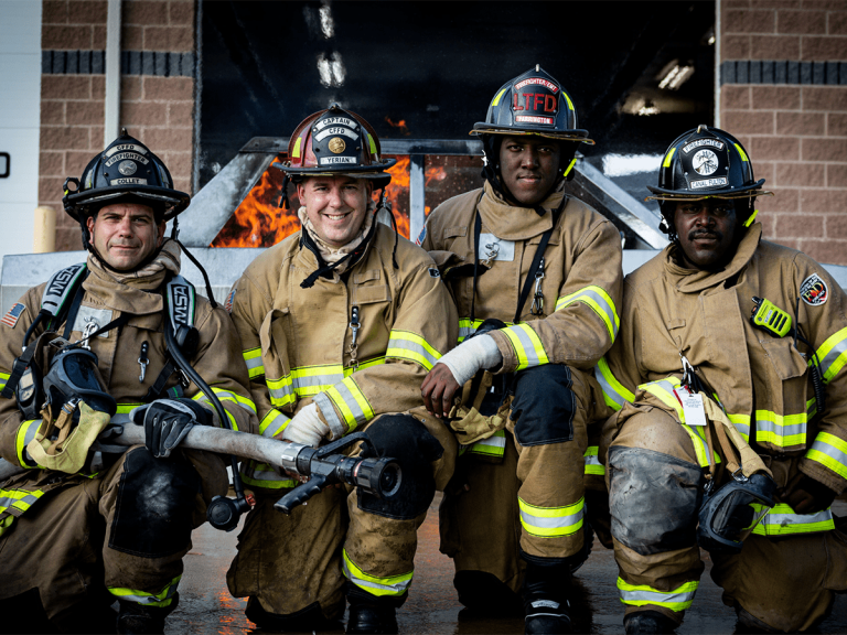 Four firefighters in full gear kneel and pose for a photo in front of a burning structure, smiling and holding fire hoses, with flames visible behind them.