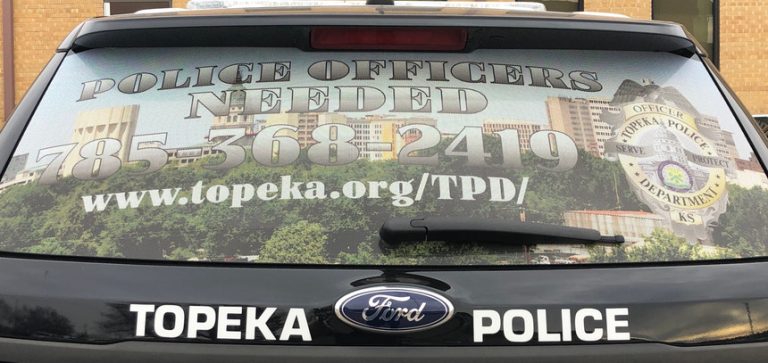 Rear window of a Topeka Police vehicle with a recruitment ad reading “Police Officers Needed,” a phone number, website link, and the Topeka Police Department badge over a cityscape background.