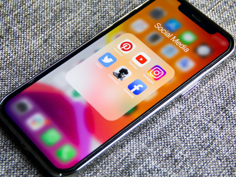 A smartphone resting on a gray fabric surface displays a folder labeled "Social Media" with Pinterest, YouTube, Instagram, Twitter, Clubhouse, and Facebook app icons on its screen.