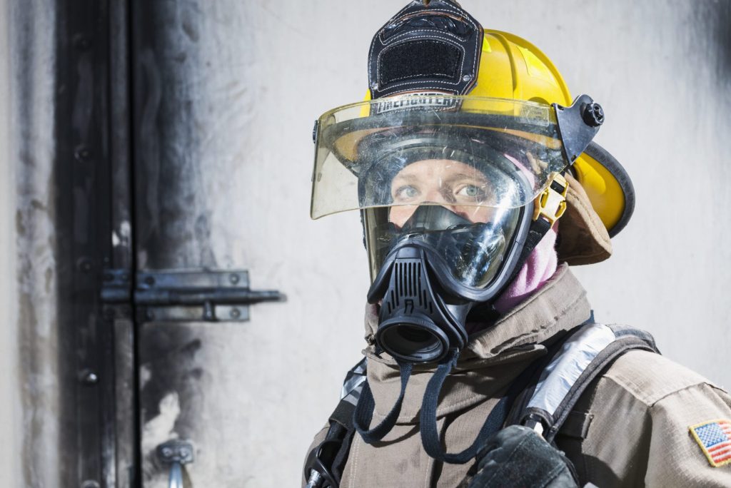 A firefighter wearing a yellow helmet, face mask, and protective gear stands in front of a dark, metal door. Sunlight highlights the firefighter’s eyes through the clear visor. An American flag patch is visible on their sleeve.