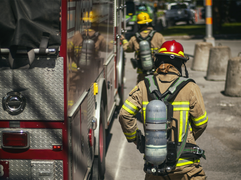 Two firefighters in full gear, including helmets and oxygen tanks, stand next to a fire truck on a sunny street with concrete barriers in the background.