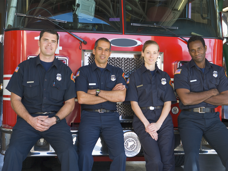 Four firefighters in uniform sit and stand in front of a red fire truck, smiling at the camera. The group includes three men and one woman, all wearing dark blue uniforms with badges.