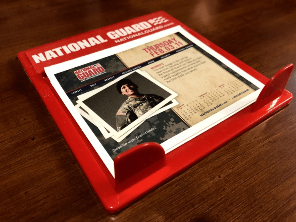 A red National Guard desk calendar sits on a wooden surface, displaying a page with a photo of a soldier in uniform and information for Thursday, February 11.