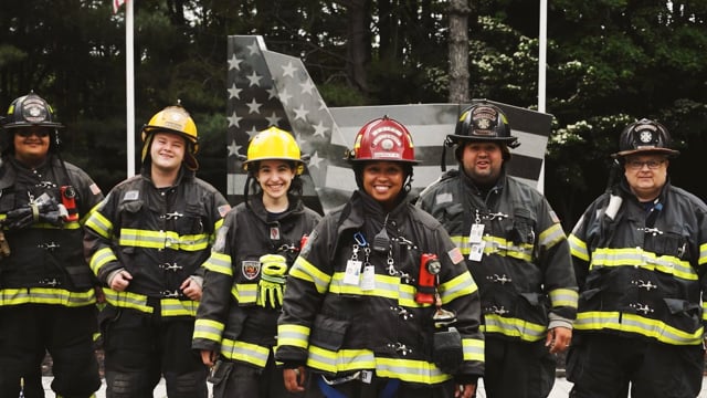 Six firefighters in full gear stand smiling in front of a black-and-white American flag mural, with trees in the background. The group includes men and women of diverse backgrounds.