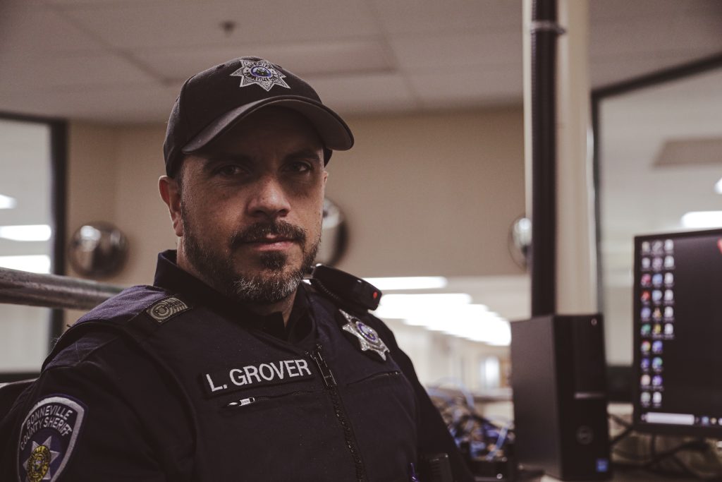 A male police officer in uniform sits indoors at a desk, looking at the camera. His badge reads "L. Grover" and he is wearing a dark cap with an emblem. There are computer monitors and office lights in the background.