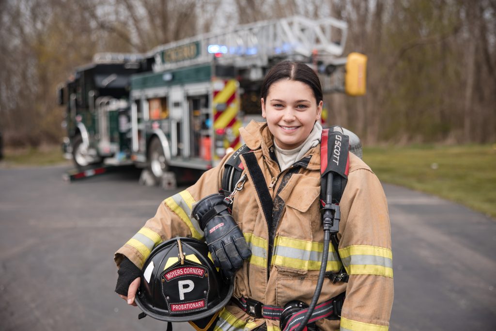 A firefighter in full gear smiles at the camera, holding a helmet labeled "WORKERS CORNERS PROBATIONARY." A fire truck with lights on is parked in the background on a paved area with trees.