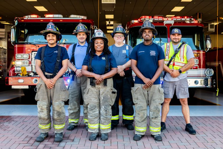 Six firefighters in uniform and helmets stand smiling in front of two red fire trucks parked inside a fire station garage. The group includes both men and women, posed on a brick driveway.