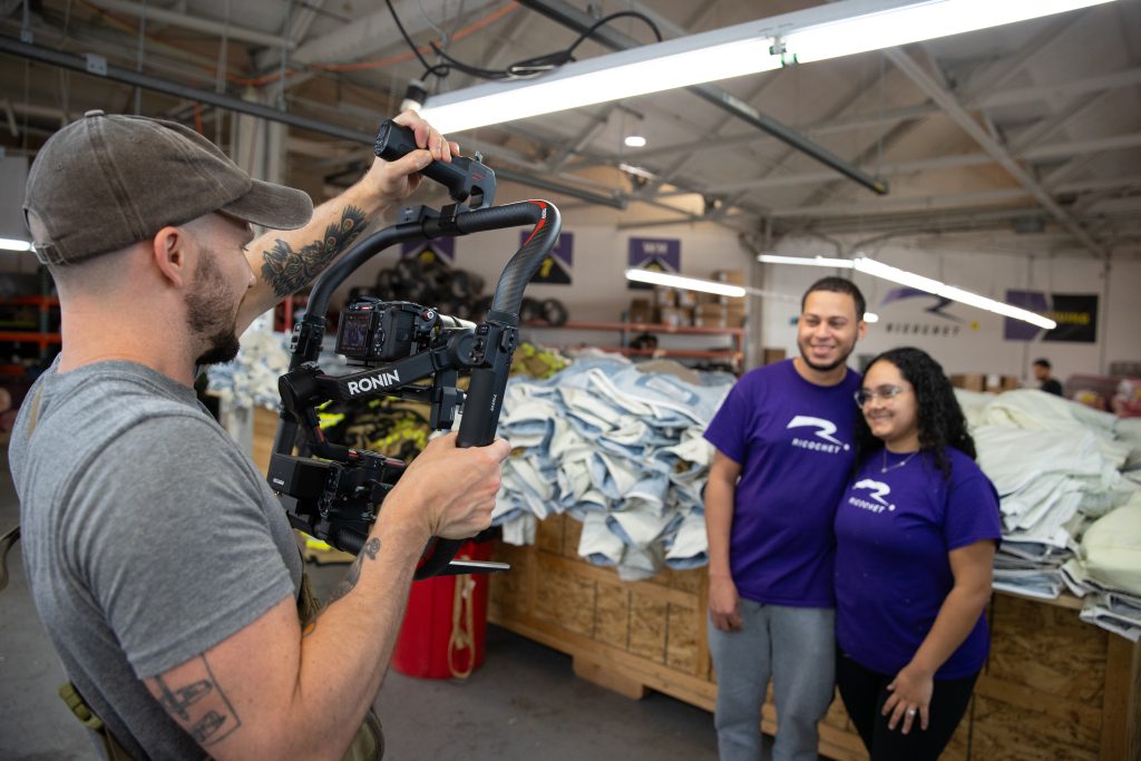 A man films a smiling couple wearing matching purple shirts in a warehouse filled with stacked fabrics and bright overhead lights.