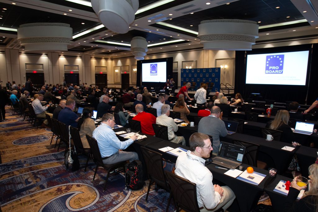 A large conference room filled with people seated at tables, facing two projector screens displaying the "Pro Board" logo. Attendees are using laptops, taking notes, and conversing. The room is well-lit and spacious.