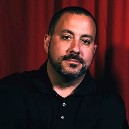 A man with short dark hair and a trimmed beard, wearing a black polo shirt, sits in front of a red curtain, looking at the camera with a neutral expression.