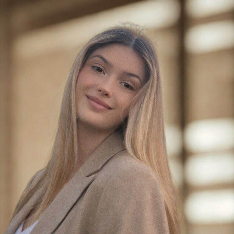 A young woman with long, straight blonde hair smiles gently at the camera. She is wearing a beige blazer and standing in front of a softly blurred background with warm tones.