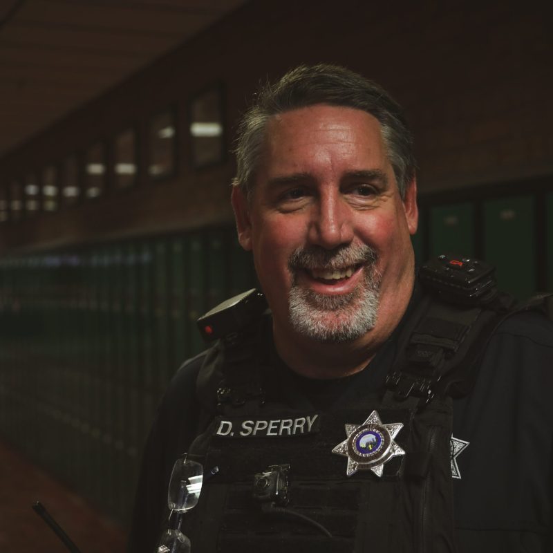 A smiling police officer in uniform stands in a hallway lined with green lockers. The officer has gray hair and beard, and his name badge reads "D. SPERRY." The lighting is dim, with the background slightly out of focus.