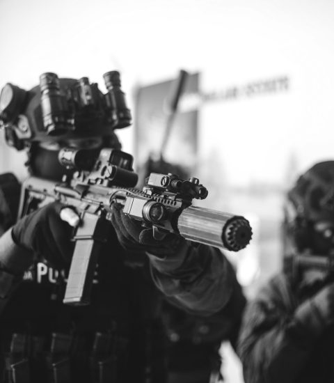Two heavily armed police officers in tactical gear and night vision goggles aim their rifles indoors, focusing on a target out of view. The image is in black and white.