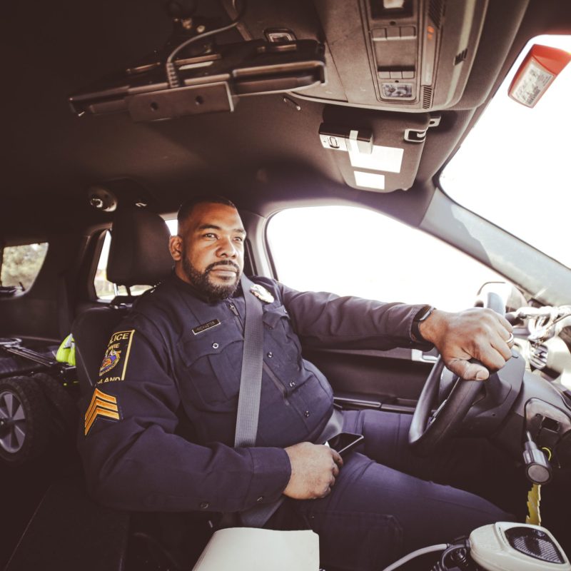 A police officer in uniform sits inside a patrol car, holding the steering wheel with one hand and a phone in the other, looking out the window. The interior of the vehicle and police equipment are visible.