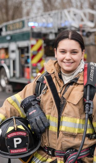 A firefighter in full gear smiles at the camera, holding a helmet labeled "WORKERS CORNERS PROBATIONARY." A fire truck with lights on is parked in the background on a paved area with trees.