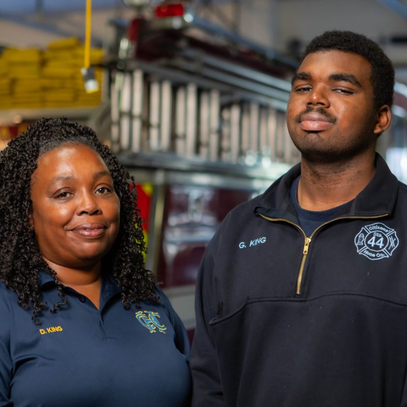 Two people in dark blue uniforms stand in front of a fire truck inside a fire station, smiling gently at the camera. The background shows firefighting equipment and the rear of the fire truck.