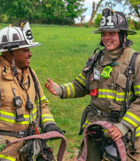 Two firefighters in full gear stand outside on grass, smiling and talking. One holds a fire hose while the other gestures with his hand. A third person is partially visible on the right. Trees and greenery are in the background.
