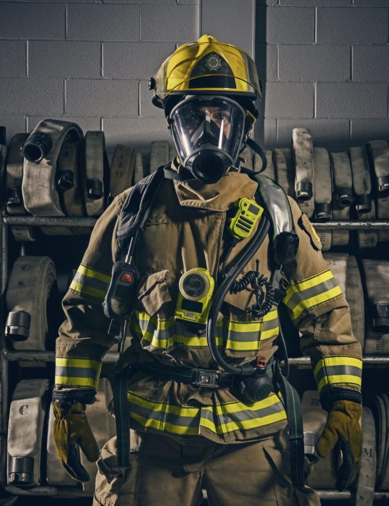 A firefighter in full protective gear, including a helmet and face mask, stands indoors in front of shelves stacked with rolled fire hoses.