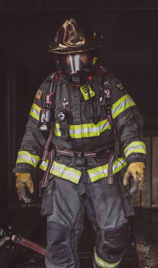A firefighter wearing full protective gear, including a helmet, gloves, and breathing apparatus, stands in a dimly lit, smoky area with a fire hose on the floor beside them.