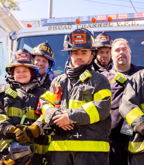 Five firefighters in full gear stand in front of a fire truck on a sunny day, posing and smiling for the camera. The fire truck behind them displays "Broad Channel V.F.D." on its sign.