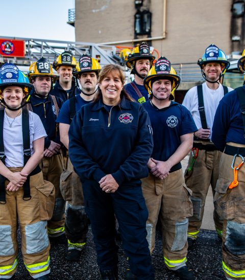 A group of firefighters in uniform and gear stand together outside, smiling at the camera. A fire truck labeled "Odenton Volunteers" is visible in the background.