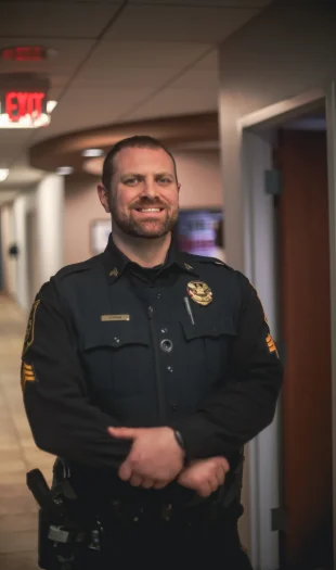 A smiling police officer in uniform stands with arms crossed in a hallway with offices and an exit sign visible in the background.