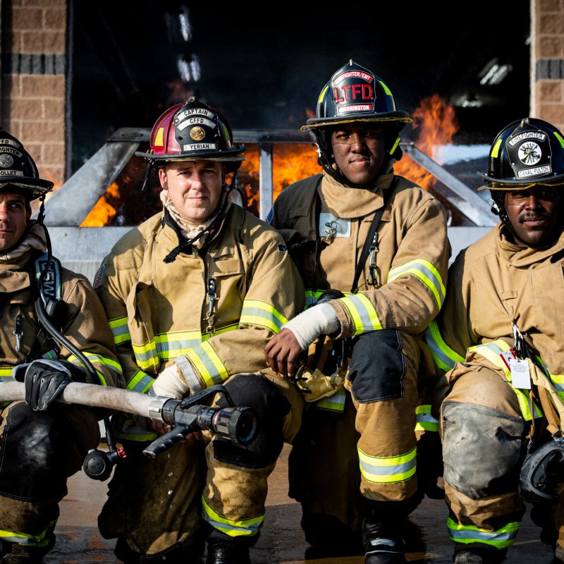 Four firefighters in full gear pose in front of a burning structure, with flames visible behind them. They are kneeling and sitting together, holding firefighting equipment, looking at the camera.